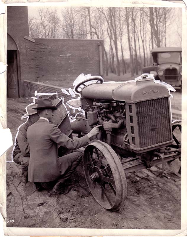 Two men kneel by a tractor they are repairing, circa 1920.